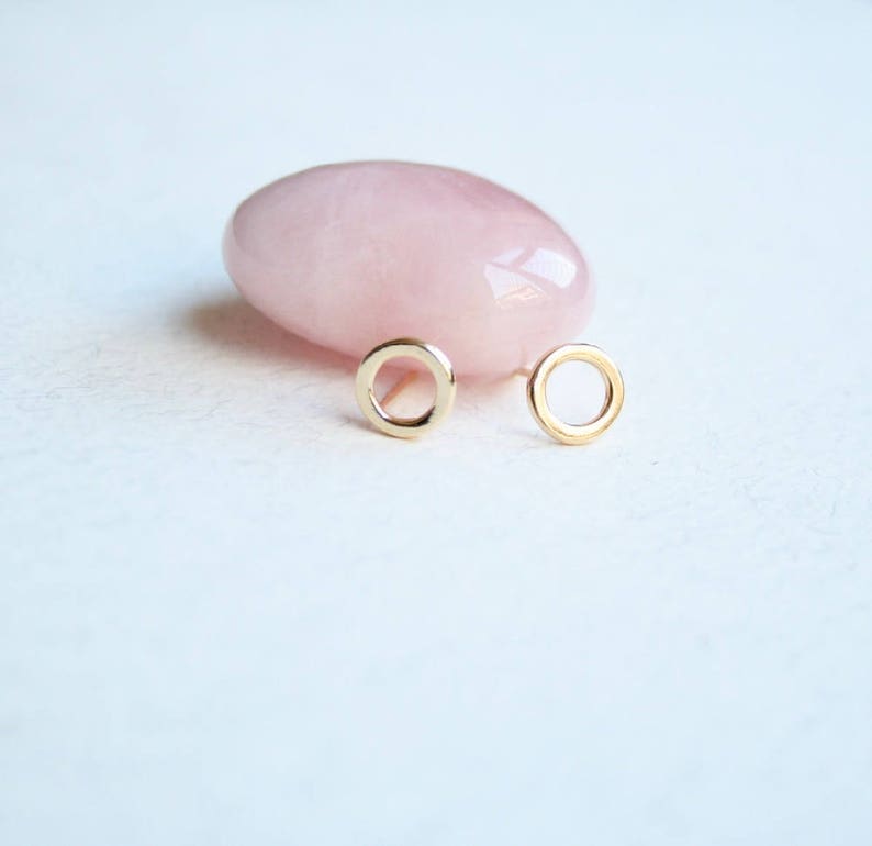 May include: A pair of small, circular gold stud earrings featuring a simple open-ring design. The earrings are positioned in front of a smooth, polished, pale pink rose quartz stone, highlighting the contrast between the metallic gold and the stone.