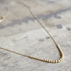 Pu&ograve; includere: Una collana color oro caratterizzata da una catenina sottile e una sezione centrale composta da piccole perline metalliche rotonde che aumentano gradualmente di dimensioni verso il centro, creando un punto focale curvo lungo la catena.