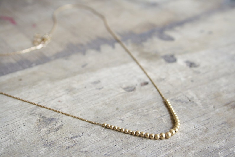 Pu&ograve; includere: Una collana color oro caratterizzata da una catenina sottile e una sezione centrale composta da piccole perline metalliche rotonde che aumentano gradualmente di dimensioni verso il centro, creando un punto focale curvo lungo la catena.
