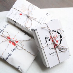 Pu&ograve; includere: Tre pacchetti rettangolari in carta bianca legati con uno spago sottile marrone e bianco. Ogni pacchetto presenta testo stampato in nero tra cui "Thank You" e "Sharon Tacker soft urban jewelry" insieme a un logo circolare e accenti rossi.