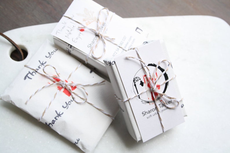 Pu&ograve; includere: Tre pacchetti rettangolari in carta bianca legati con uno spago sottile marrone e bianco. Ogni pacchetto presenta testo stampato in nero tra cui "Thank You" e "Sharon Tacker soft urban jewelry" insieme a un logo circolare e accenti rossi.