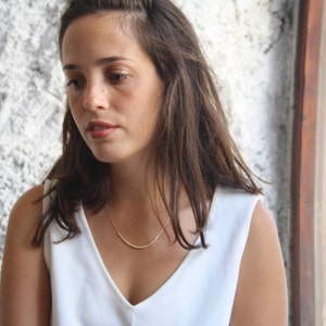 Pu&ograve; includere: Una delicata collana color oro con un ciondolo a barra curva caratterizzato da una superficie testurizzata a perline. La collana &egrave; indossata con un semplice top bianco senza maniche, che mette in risalto il design minimalista del gioiello.