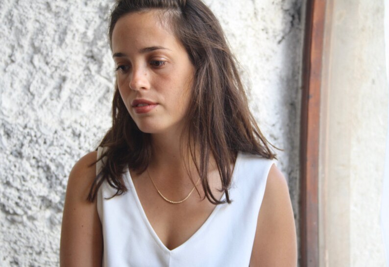 Pu&ograve; includere: Una delicata collana color oro con un ciondolo a barra curva caratterizzato da una superficie testurizzata a perline. La collana &egrave; indossata con un semplice top bianco senza maniche, che mette in risalto il design minimalista del gioiello.
