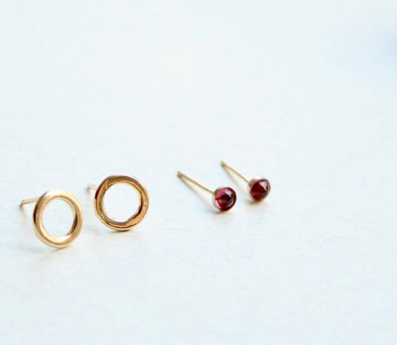 May include: Two pairs of stud earrings. One pair features simple gold-toned circular hoops, while the other pair consists of small, round, deep red gemstone studs set in gold-toned metal. Each earring has a thin post for secure attachment.