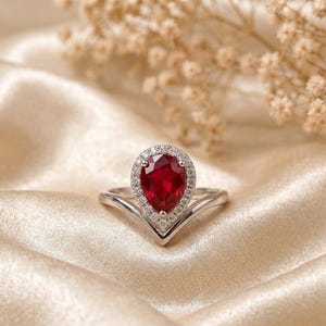 May include: A silver-toned ring featuring a pear-shaped red gemstone as the centre stone, surrounded by a halo of small white stones. The band has a V-shaped design that meets at the base of the pear-shaped setting, creating a pointed silhouette.