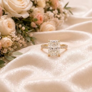 Op de afbeelding: Een gouden ring met een grote rechthoekige radiant-geslepen centrale steen en een pavé-gezette band. De ring ligt op een zachte, gedrapeerde crèmekleurige stof, met een boeket van lichtroze en witte rozen en groen blad op de achtergrond.