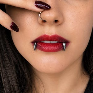 May include: A pair of silver-toned metal fang-shaped lip piercings positioned on the lower lip. The jewelry features a smooth, polished finish and a curved, pointed design that mimics the appearance of vampire fangs resting against the teeth.