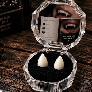 May include: A pair of white vampire fangs resting in a black velvet-lined, clear octagonal plastic case. The lid features a mirror with a printed instruction card titled HOW TO USE, listing three steps and the text CONFIDENCE LIVES HERE.