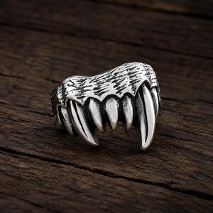 May include: A silver-toned metal ring featuring a sculpted design of a row of sharp, pointed animal teeth or fangs. The surface of the ring has a textured, dark oxidized finish in the recessed areas to highlight the detailed shape of the fangs.