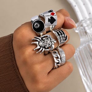 May include: A set of silver-toned rings featuring various designs including a black eight ball, playing card suits, a skeleton hand, a jester face, a thick chain link band, and a band with small playing card symbols engraved on the surface.