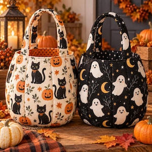 Puede incluir: Dos cestas de tela acolchada con asas. La cesta de la izquierda es de color crema con un estampado de gatos negros, calabazas de Halloween naranjas y hojas. La cesta de la derecha es negra con un estampado de fantasmas blancos, lunas y estrellas.