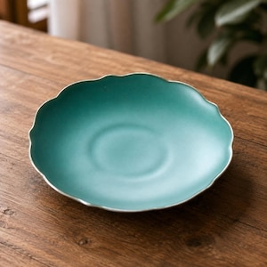 Könnte beinhalten: Eine runde, türkisfarbene Untertasse aus Keramik mit einem gewellten, muschelförmigen Rand. Der Teller hat eine glatte, matte Oberfläche und eine leicht vertiefte kreisförmige Mitte, die für eine Tasse vorgesehen ist. Der Rand ist dünn und geschwungen.