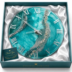 May include: A round wall clock with a teal and white marbled resin face, silver Roman numerals, and silver hands. A textured band of silver glitter and small beads runs diagonally across the center. A card reads Thank You FOR YOUR PURCHASE.