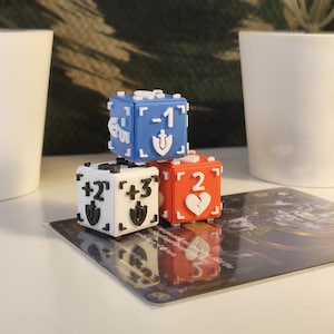 Peut inclure: Trois dés de jeu à six faces empilés. Le dé supérieur est bleu avec un -1 blanc et une icône de bouclier. Le dé en bas à gauche est blanc avec des marquages +2 et +3 noirs et des icônes de bouclier. Le dé rouge a un 2 blanc et un cœur.