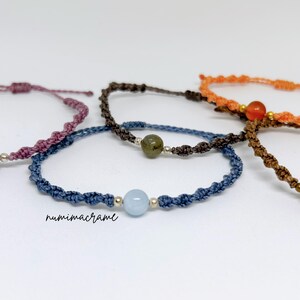 May include: Five macrame bracelets featuring twisted cord bands in purple, dark brown, orange, blue, and brown. Each bracelet includes a single central gemstone bead flanked by small metallic spacers. The text numimacrame is visible in black.