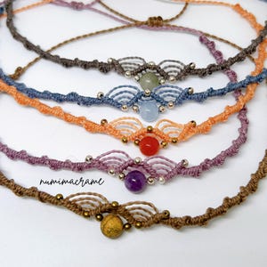 May include: Five macrame bracelets featuring knotted cord in brown, purple, orange, blue, and grey. Each bracelet includes a central gemstone bead, metallic accents, and a decorative arched knot pattern. The text numimacrame is visible on the left.