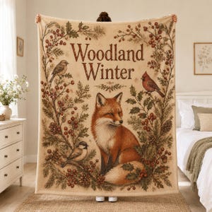 Könnte beinhalten: Eine cremefarbene Wohndecke mit dem Schriftzug Woodland Winter in brauner Schreibschrift. Das Design zeigt einen Fuchs in der Mitte, umgeben von Tannenzweigen, roten Beeren, Tannenzapfen und Vögeln, darunter ein Kardinal und ein kleiner Singvogel.