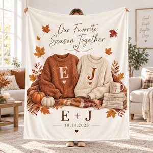 Può includere: Una coperta color crema con il testo Our Favorite Season Together, due maglioni a trecce con le iniziali E e J, una pila di libri intitolati Pumpkin, Sweaters, Good Company, Brighter Days, zucche e la data 14.10.2023.