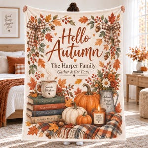 Op de afbeelding: Een crèmekleurige plaid met de tekst Hello Autumn, The Harper Family en Gather & Get Cozy. Het ontwerp bevat herfstbladeren, eikels, geruite strikken, een stapel boeken, pompoenen, een mok, een kaars en een geruite deken.
