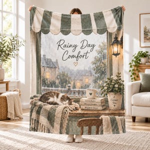 Puede incluir: Una manta suave con un estampado de un gato durmiendo en el alféizar de una ventana, libros apilados, una planta en maceta y un farol. El texto dice "Rainy Day Comfort" con un corazón, "Good Books", "Warm Drinks", "Brighter Days" y "A Calmer You".