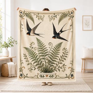 Puede incluir: Una manta de color crema con una ilustración central de frondas de helecho verdes y dos golondrinas volando con marcas azul oscuro y naranja. El diseño está encerrado en un borde decorativo con pequeñas flores azules y un icono de corazón.