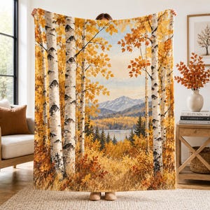May include: A rectangular throw blanket featuring a printed landscape scene of a forest with white birch trees in the foreground, golden autumn foliage, a calm lake, and distant mountains under a pale sky, rendered in warm yellow and orange tones.