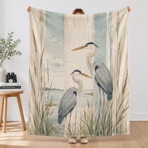 Può includere: Una coperta rettangolare con un'illustrazione stampata di due aironi cenerini in piedi tra canne alte. Lo sfondo mostra una scena costiera dai toni tenui con strisce verticali color beige, crema e blu tenue, con due uccelli in volo.