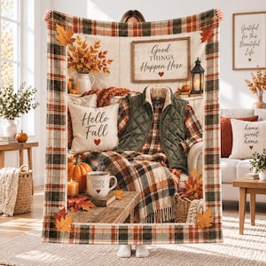 Könnte beinhalten: Eine rechteckige Wohndecke mit einem karierten Rand in Orange-, Grün- und Brauntönen. Die Mitte zeigt eine gemütliche Herbstszene mit einer Steppweste, einem Kissen mit der Aufschrift Hello Fall, einer Tasse mit Cozy Days und einem Schild Good Things Happen Here.