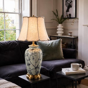 May include: A table lamp with a light green ceramic base featuring a blue bird and branch pattern, a gold metal base, and a beige fabric lampshade with a scalloped edge. The lamp emits a warm glow, illuminating the intricate botanical design.