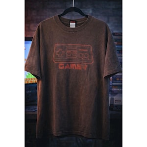 Może przedstawiać: Brązowy t-shirt z krótkim rękawem z wyblakłą czerwoną grafiką przedstawiającą zarys kontrolera do gier Nintendo oraz nadrukowanym poniżej słowem GAMER. Koszulka ma okrągły dekolt i tkaninę o spranym, postarzanym wyglądzie.