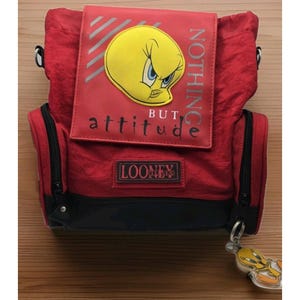May include: A red backpack featuring a yellow Tweety Bird graphic on the front flap with the text NOTHING BUT attitude and a Looney Tunes patch. The bag includes black trim, side zippered pockets, and a dangling Tweety Bird character keychain.
