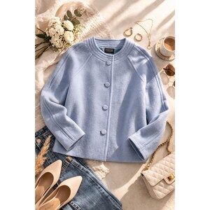 May include: A light blue, long-sleeved jacket with a round neckline and four matching buttons down the front. The jacket features a textured fabric, structured shoulders, and a Pendleton brand label on the inside collar. The garment is displayed alongside blue jeans, beige pointed-toe shoes, a white quilted handbag, gold jewellery, and a small bouquet of white flowers.