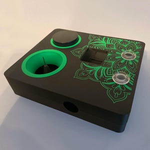 May include: A black rectangular electronic device featuring a bright green circular recessed area with a central pin, a solid black circular button, a square cutout, and two small clear circular buttons. A green mandala pattern is printed on the top.