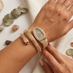 May include: A gold-toned watch featuring a snake-inspired design with a flexible, segmented band that wraps around the wrist. The watch face is oval-shaped, bordered by small clear stones, and displays a textured, patterned dial with gold hands.