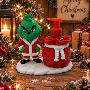Puede incluir: Una figura verde del Grinch con traje de Papá Noel rojo y blanco junto a un dispensador de jabón rojo con forma de saco y cierre de cuerda blanca, sobre una base blanca que simula nieve, con un cabezal de bomba para el jabón líquido.