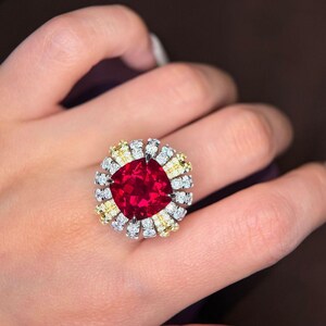 Puede incluir: Un anillo con una gran piedra central roja de corte cojín rodeada por un halo de piedras redondas alternadas en blanco y amarillo. El engaste es metálico y el diseño crea una disposición circular similar a una flor.