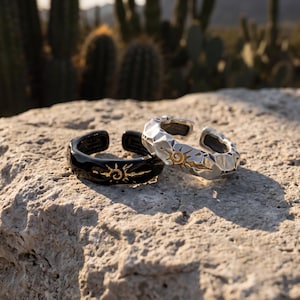 May include: Two open-band rings, one in a glossy black finish and the other in a textured silver finish. Both rings feature a central gold-colored sunburst engraving with a spiral center, showcasing a bold, geometric design on the outer surface.