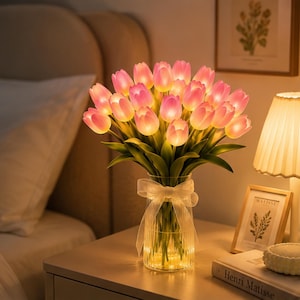 May include: A bouquet of artificial pink tulip flowers with integrated LED lights, arranged in a clear, vertically ribbed glass vase. The vase is decorated with a sheer white ribbon tied in a bow around the neck, and the tulips emit a soft glow.