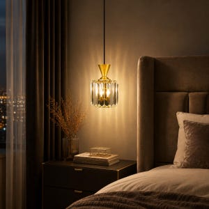 May include: A pendant light fixture featuring a cylindrical, faceted glass shade with a gold-toned metal top and a thin black cord. The light emits a warm glow, casting vertical light patterns onto the wall behind it, creating a bright illumination.
