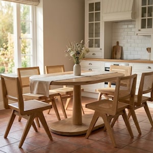 May include: A light oak dining set featuring an oval table with a central pedestal base and six chairs with woven cane backrests and seats. A beige fabric table runner lies across the centre of the table, topped with a white vase of dried flowers.