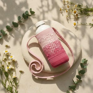 Puede incluir: Una botella de agua blanca envuelta en una funda de ganchillo con un degradado de color que va del rosa claro en la parte superior al rosa intenso en la base. La funda incluye una correa larga de ganchillo a juego para transportarla.