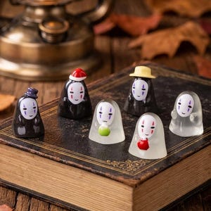 May include: Five small figurines of the character No-Face from Spirited Away, each with a different accessory including a hat, a red cap, a yellow hat, a green ball, and a red apple, arranged on the cover of a vintage, leather-bound book.