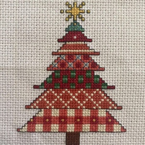 May include: A cross-stitch Christmas tree design. The tree has red, green, and white patterns, including a plaid base and a star on top. The tree is stitched on a light beige fabric.