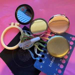 May include: A set of embroidery supplies including a wooden embroidery hoop, a black fabric square, a needle threader, a variety of embroidery floss, a small round mirror, a larger round mirror with a gold finish, and a blue plastic embroidery thread organiser with numbered holes.
