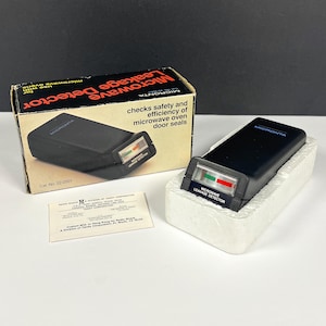 May include: A black Micronata microwave leakage detector with a green and red indicator gauge, housed in a white styrofoam tray, accompanied by its original cardboard box featuring product text and a small informational paper insert.