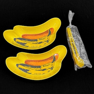 May include: Two yellow banana-shaped bowls featuring a graphic print of a banana with the signature Andy Warhol. The set includes a pair of yellow plastic tongs sealed in a clear plastic bag. The bowls have a curved design and bright yellow finish.