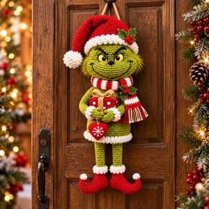 May include: A crocheted green character wall hanging wearing a red Santa hat with a white pom-pom, a red and white striped scarf, and red elf shoes. The figure holds a red gift box with white polka dots and a small red heart ornament.