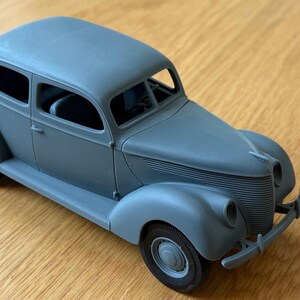Könnte beinhalten: Ein mattgraues Modellauto einer alten Limousine mit abgerundeter Karosserie, markanten vorderen Kotflügeln, einem horizontalen Lamellengrill, runden Scheinwerfern und vier Rädern. Der Innenraum ist durch die Fensteröffnungen sichtbar.