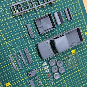 Op de afbeelding: Een grijze plastic modelvrachtwagenkit met een chassis, laadbak, cabine, vier wielen en diverse kleine onderdelen op een groene snijmat. De set bevat een voorruit van doorzichtig plastic en diverse ongeverfde grijze onderdelen.