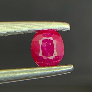 May include: A vibrant, oval-cut ruby gemstone with a deep red hue and multifaceted surface, held securely between the two metallic prongs of a pair of silver-toned tweezers, showcasing its polished clarity and distinct geometric faceting.